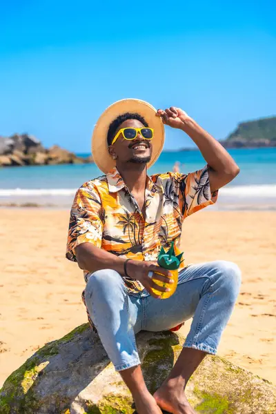 Güneş gözlüğü ve hasır şapka takan genç adam güneşli bir plajda bir kayanın üzerinde dinlenirken ananas şeklinde bir kokteyl tutuyor.