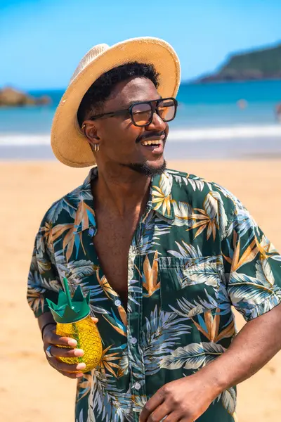 Şık güneş gözlüğü ve modaya uygun bir şapka takan genç adam güneşli bir sahilde dinlenirken ferahlatıcı bir kokteylin tadını çıkarıyor.