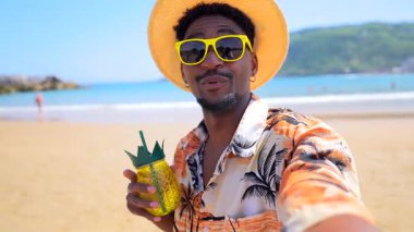 Güneş gözlüğü takan ve hasır şapkalı genç adam güneşli bir sahilde ferahlatıcı bir ananas kokteylinin tadını çıkarıyor.