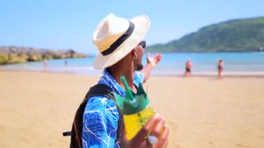 Genç siyah adam güneş gözlüğü takıyor ve şık hasır şapka takıyor. Taze ananas kokteyli ile rahatlıyor. Okyanus manzaralı güneşli tropikal plaj sahnesinin ve kaygısız yaz atmosferinin tadını çıkarıyor.