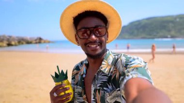 Neşeli siyah adam kumsalda dinleniyor, tatil anını ananas şekilli kokteyl ve moda yaz tarzıyla yakalıyor.