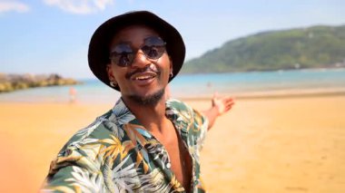 Siyah erkek içerik yaratıcısı güneş gözlüğü takıyor ve kişisel video bloğunu kaydediyor. Okyanus dalgaları ve mavi gökyüzü olan tropik plajda duruyor.