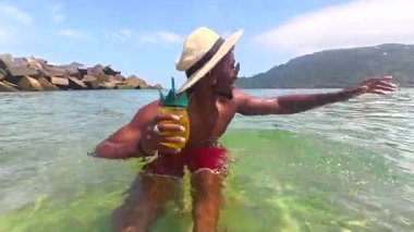 Meyveli ananas kokteyli yudumlayan genç gezgin berrak tropikal okyanus sularında güneş gözlüğü ve yaz şapkası takarak dururken neşe saçıyor.
