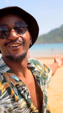 Neşeli genç siyah adam tropikal plaj tatilinin tadını çıkarıyor, güneş gözlüğü ve kova şapka takıyor, yaz neşesi ve rahatlığı yayıyor.