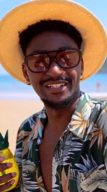 Güneş gözlüklü ve hasır şapkalı genç siyah adam sahilde ananas şeklinde bir kokteyl ile poz veriyor.