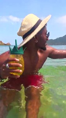 Güneş gözlüğü ve hasır şapka takan genç siyah adam berrak tropikal deniz suyunda ananas kokteylinin tadını çıkarıyor.