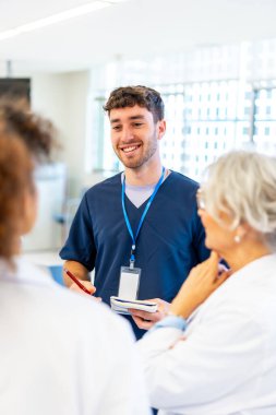 Hastane koridorunda tıp ekibiyle konuşurken gülümseyen ve not alan genç erkek hemşire.