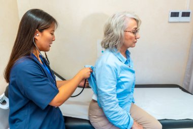 Klinikteki rutin bir muayenede steteskopla yaşlı bir kadının akciğerlerini inceleyen kadın doktor.
