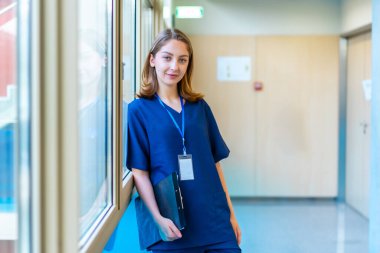 Modern bir hastane koridorunda dururken mavi önlük giymiş, elinde bir pano tutan kendinden emin genç bir doktor.