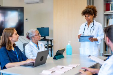 Tıbbi brifinge katılan doktorlar ve hemşireler, modern bir hastane ortamında hasta verilerini ve tedavi planlarını analiz ediyorlar.