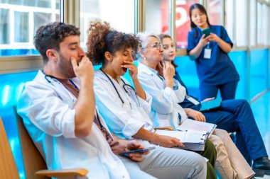 Hastane koridorunda sandalyelerde oturup uzun bir vardiyadan sonra yorgunluk çeken yorgun tıbbi uzmanlar esniyorlar.