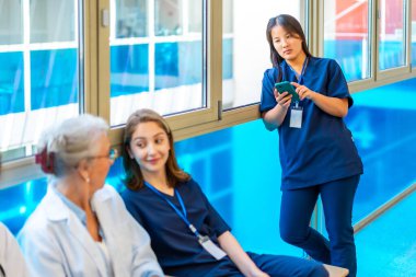 Meslektaşları hastane koridorunda konuşurken Asyalı hemşire akıllı telefon kullanıyor ve dinleniyor. Takım çalışmasının keyfini çıkarıyor.