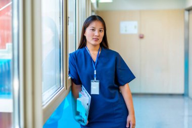 Kendine güvenen genç doktor elinde bir pano ve mavi önlükle hastane koridorunda bir pencerenin yanında duruyor.