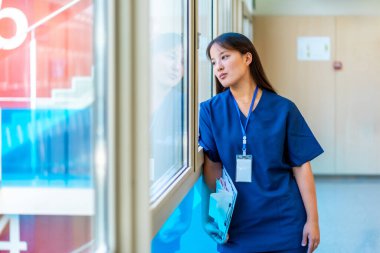 Hastane koridorunda durup, hasta dosyalarını tutarak, düşünceli bir ifadeyle dışarıyı seyreden düşünceli bir tıbbi uzman.