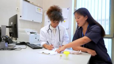 Biri beyaz önlüklü, diğeri önlüklü iki kadın doktor, hastane ofisinde birlikte tıbbi kayıtları incelerler.