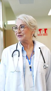 Hastane koridorunda kendine güvenen, beyaz laboratuvar önlüğü ve steteskop giyen, tıbbi uzmanlık ve deneyimi temsil eden profesyonel kıdemli kadın doktor.