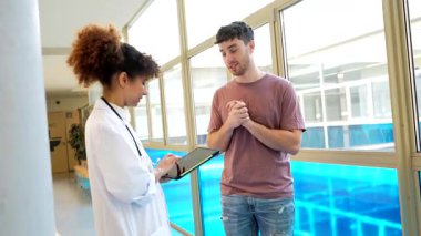 Parlak hastane koridorunda tıbbi danışma sırasında hasta bilgilerini belgeleyen kadın sağlık uzmanı, kapsamlı bakım etkileşimi için dijital tablet kullanıyor.