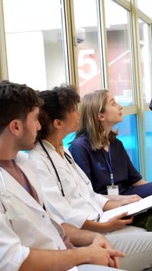 Beyaz üniformalı tıp uzmanları, hastane koridorunda yürürken hasta bilgilerini paylaşıyorlar, sağlık durumlarını tartışıyorlar.