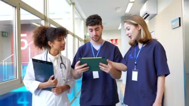 Hastane koridorunda yürürken iş birliği yapan tıp uzmanları tabletteki dijital sağlık kayıtlarını inceliyor, hasta bilgilerini odaklanmış uzmanlıkla tartışıyorlar.