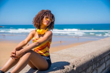 Genç bir kadın yaz güneşi ve okyanus esintisini emiyor, plajda dinlenirken kulaklıklarıyla müziğin tadını çıkarıyor.