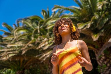 Canlı bir tropikal cennette kulaklık takan turist, güneşli mavi gökyüzünün altında sallanan palmiyelerle çevrili.