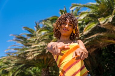 Kulaklık takan zarif genç bir kadın tropik bir yerde palmiye ağaçlarının önündeki kamerayı işaret ediyor.