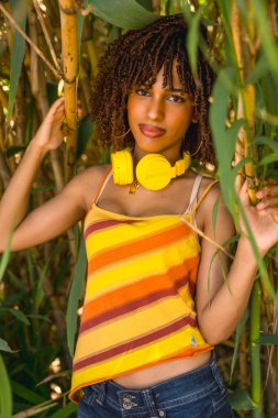 Sarı kulaklıklı genç bir kadın bambu ormanında müzik dinliyor, renkli çizgili bir bluz ve kot pantolon giyiyor.