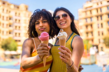 Güneşli bir yaz gününde, canlı bir şehirde renkli dondurma külahlarını tutarken gülümseyen iki genç kadın birlikte vakit geçirmenin keyfini çıkarıyorlar.