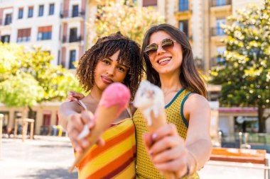 İki arkadaş şehirde güneşli bir günün tadını çıkarıyorlar. Sıcak güneş ışığında gülümserken renkli dondurma külahlarını kucaklayıp paylaşıyorlar.