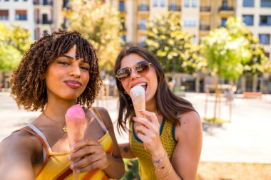 İki neşeli kadın açık havada dondurma yiyor güneşli bir yaz gününde şehir ortamında selfie çekiyorlar.