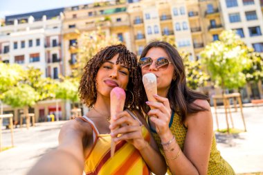 İki arkadaş canlı bir şehir meydanında güneşli bir günde dondurma külahlarının tadını çıkarıyor. Şehir ortamında neşe ve dostluk sergiliyorlar.