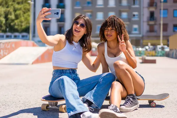 İki genç kadın, kaykay parkında güneşli bir günde kaykay yaparken akıllı telefonlarıyla selfie çekiyorlar.
