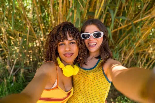 İki gülümseyen kız bambu bitkilerinin önünde selfie çekiyor, sarı elbiseler, kulaklıklar ve güneş gözlüğü takıyorlar.