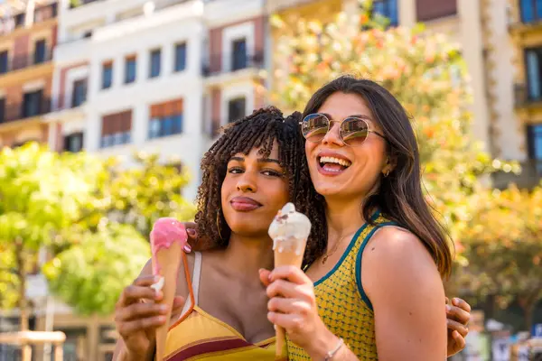 İki gülümseyen kız dondurma yiyor, güneşli bir yaz gününde şehir merkezinde birbirlerine sarılıyorlar.