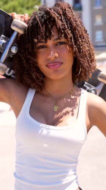 Omuzlarında kaykayla güvenle duran genç bir kadın kentli yaşam tarzını ve kişisel tarzını göz alıcı bir gülümsemeyle sergiliyor.