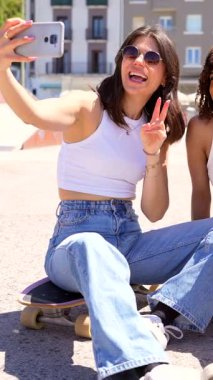 Genç kadınlar selfie çekerken kaynaşıyor, şehir ortamında kaykayın üzerinde oturuyor, arkadaşlığı ve neşeli yaz anını cep telefonuyla yakalıyor.