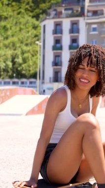 Kıvırcık saçlı genç Afro-Amerikan kadın kaykaya güvenle oturuyor, güneşli şehir kaykay pistinde gülümsüyor, günlük yaz kıyafetleri giyiyor.