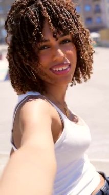 Kıvırcık saçlı genç bir kadın, altın saat güneş ışığı altında şehir manzarasını incelerken anlamlı aralıklarla selfie çekiyor.