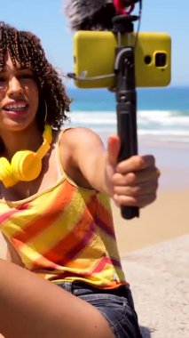 İçerik yaratıcı güneşli sahilde vlog yapıyor, dış mikrofonlu akıllı telefon kullanıyor, enerjik hikaye anlatımı ve dijital yaratıcılık ile yaşam tarzı anını yakalıyor