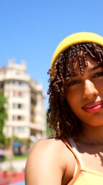 Sarı bereli ve altın kolyeli oyuncu genç bir kadın güneşli bir şehir ortamında kameraya çeşitli yüz ifadeleri takınıyor.