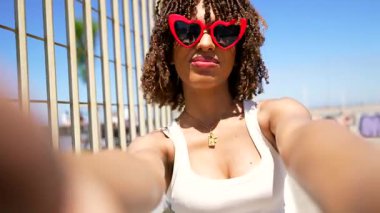 Canlı kırmızı kalp takan genç bir kadın güneş gözlüğü şeklindeki selfie anını yakalıyor metal çitlerin yanında, son moda yaz stili ve kendinden emin bir enerji yayıyor.