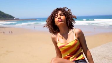 Kıvırcık saçlı, güneş ışığında güneşlenen, plaj duvarında oturan yaz tatili boyunca deniz melteminin ve huzurlu sahil atmosferinin tadını çıkaran genç bir kadın.