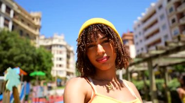 Sosyal medya içeriği yaratan, şık sarı şapka ve günlük yaz kıyafetleri giyerken şehir parkı manzarasının ortasında güvenle poz veren genç bir kadın etkili.