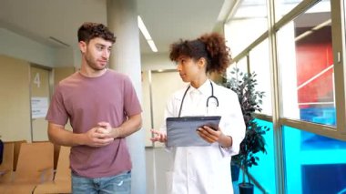 Genç doktor, hastane koridorunda yürürken hastaya tedavi seçenekleri ve sağlık hizmetleri iletişimi hakkında danışmanlık yapar.