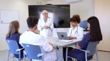 Doktorlar modern bir hastane konferans odasında hasta bakımı stratejilerini tartışıyor ve işbirliği yapıyorlar. Etkili iletişim için dizüstü bilgisayarlar ve not defterleri kullanılır.