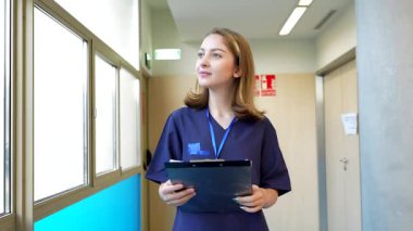 Genç bayan doktor, parlak bir hastane koridorunda yürürken hasta notlarını incelikle gözden geçirip, muayeneye hazırlanıyor.