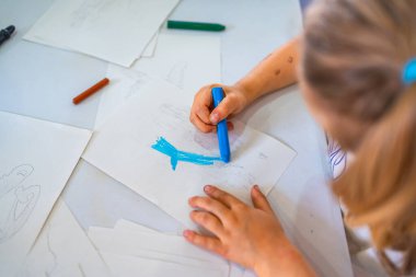 Masasındaki parlak beyaz kağıda mavi mum boya kalemi çizerken dikkatle konsantre olan genç kız yaratıcılığını gösteriyor.