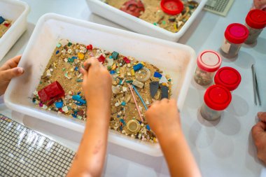 Öğrenciler bir laboratuvar deneyi sırasında beyaz plastik bir kaptaki kumdan renkli mikro plastikler topluyorlar.