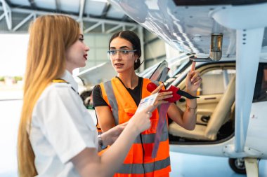 Yüksek görüş mesafesinde yelek giyen kadın uçak bakım mühendisi, uçak parçalarını hangarda belgeleri tutan pilota anlatıyor.
