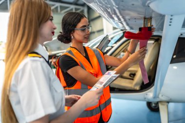 Kadın uçak bakım mühendisi pilot eşliğinde uçak kanadını teftiş ediyor, kontrol listesi tutuyor ve hangarda prosedürleri tartışıyor.